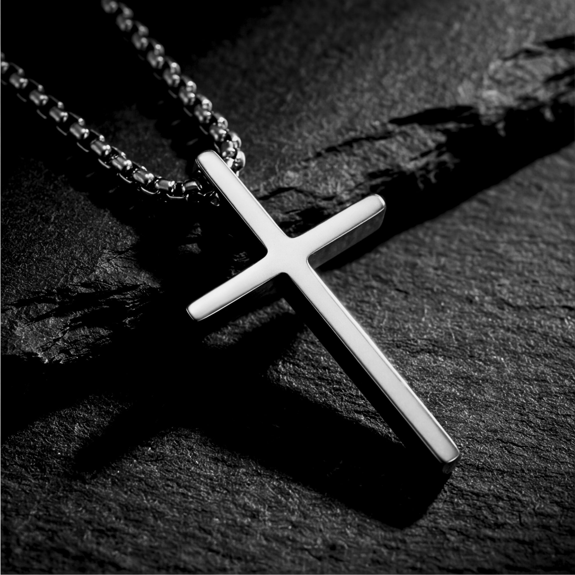 Silver stainless steel cross pendant on a box chain, resting on a dark textured stone surface.