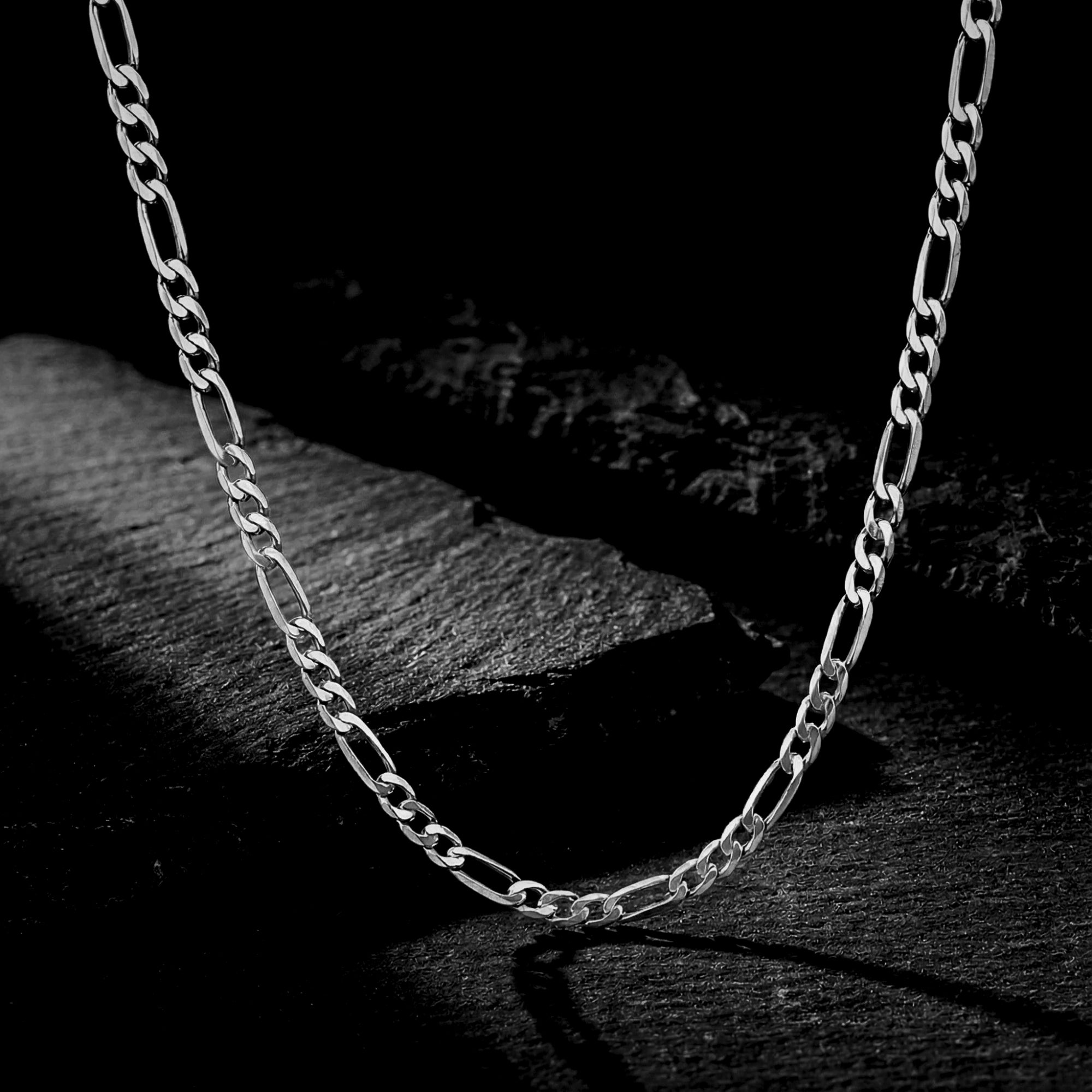 Silver 3mm stainless steel Figaro chain draped over dark textured rocks.
