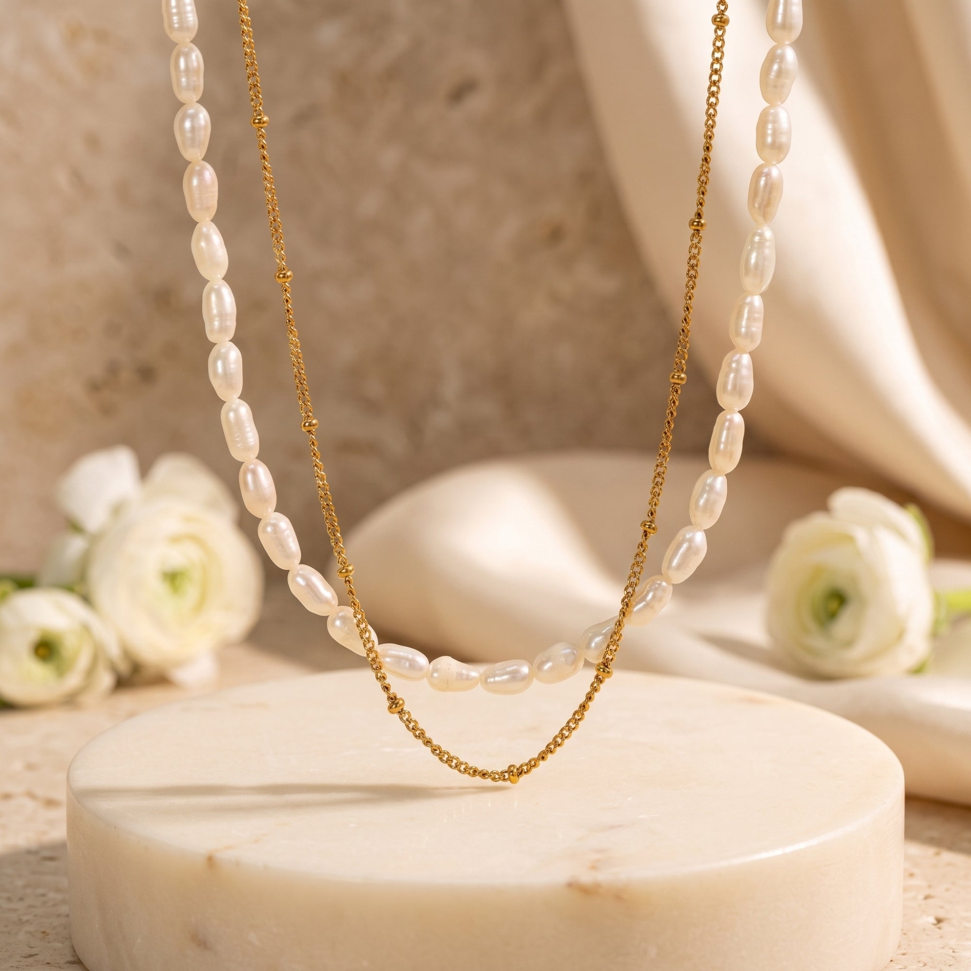 Layered gold satellite chain and freshwater pearl necklace on a stone pedestal with white flowers.