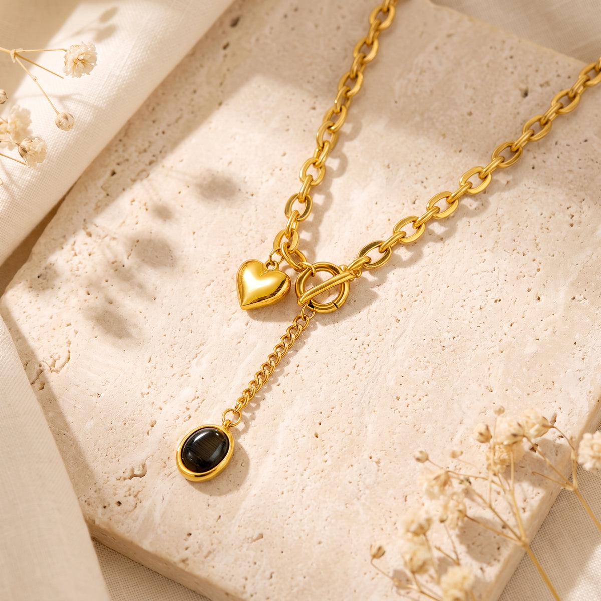 Gold-tone heart lariat necklace with a dark oval pendant arranged on a beige stone surface with dried flowers.