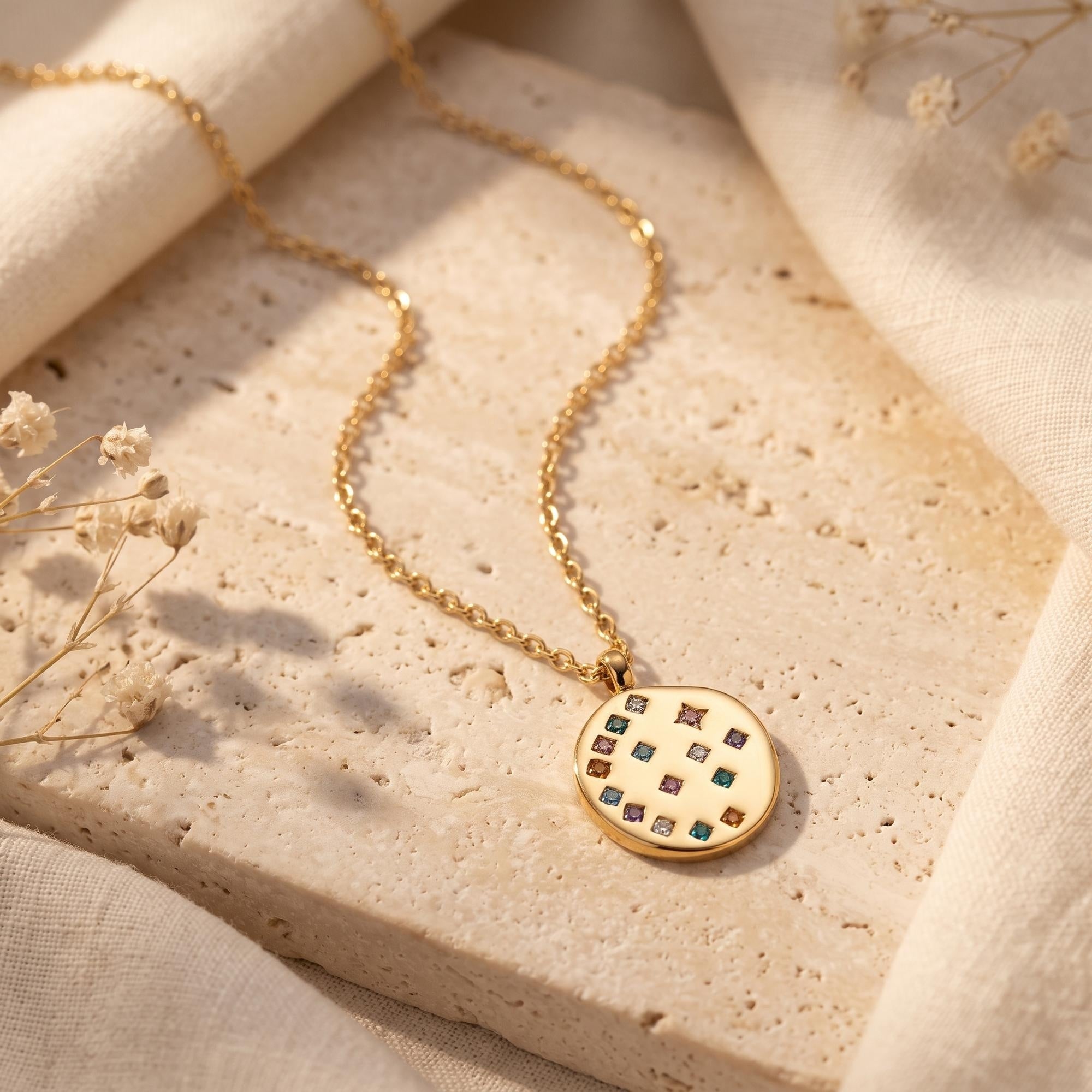 18K gold Astraline circular medallion necklace with multi-color CZ stones on a stone surface with dried flowers.