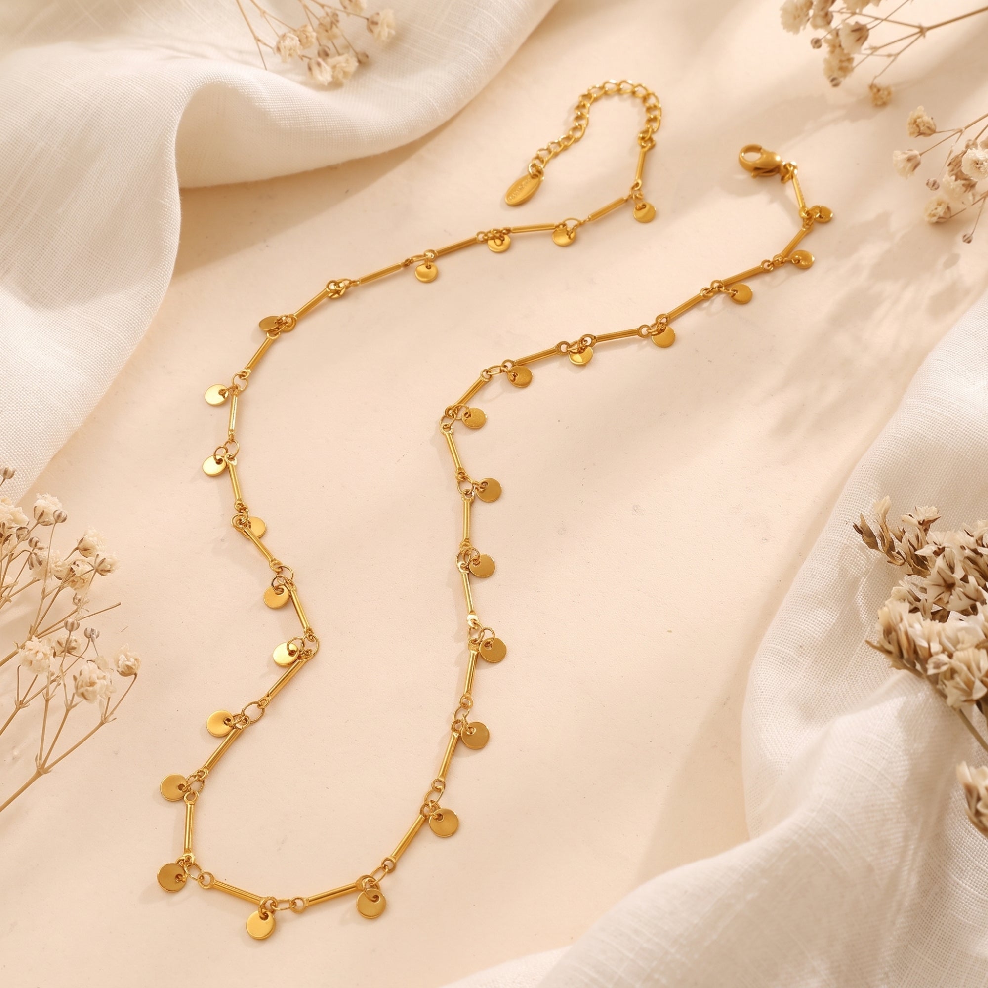 Gold disc necklace on white fabric with dried flowers.