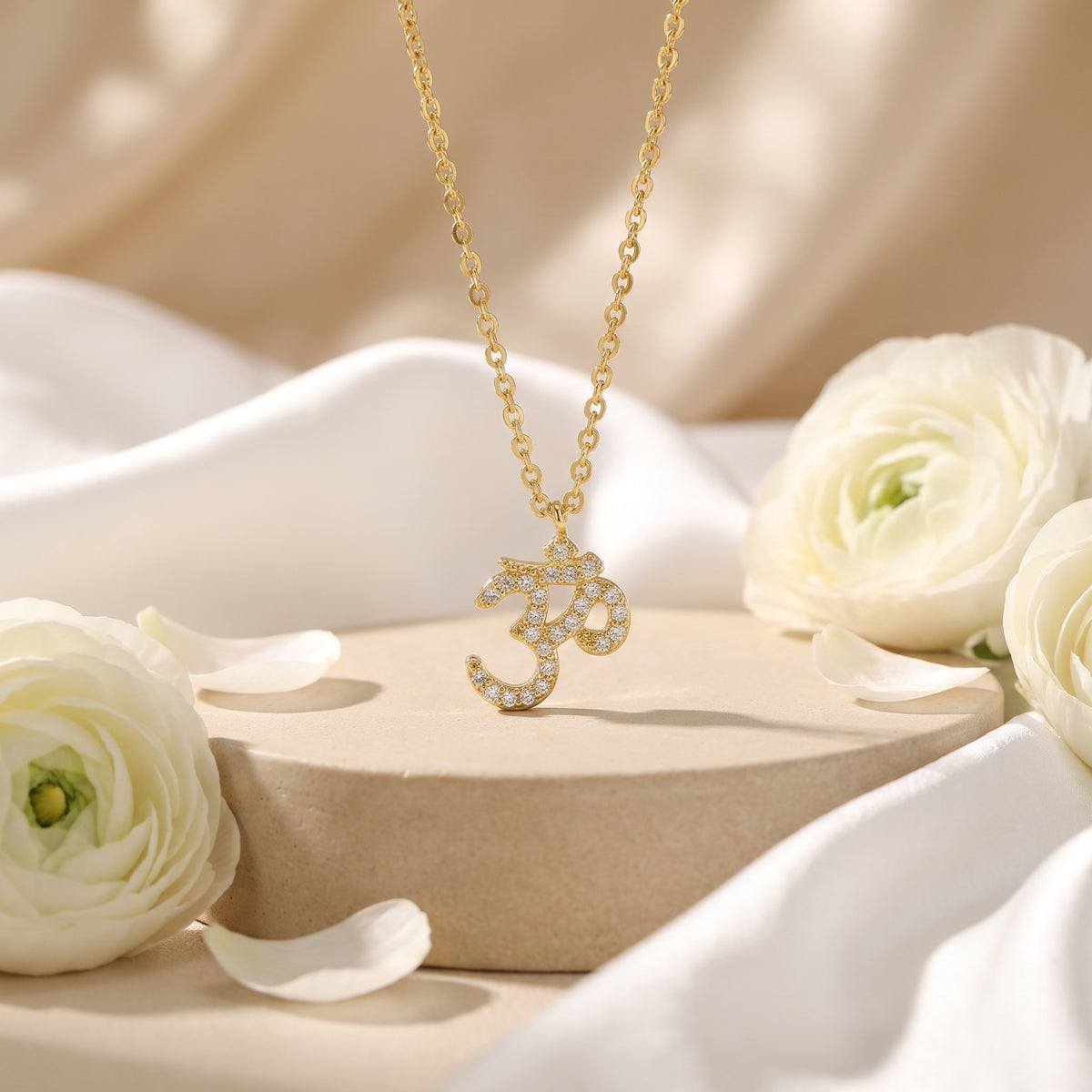 Gold-tone Om pendant necklace with clear stone accents, displayed on a podium with white flowers.
