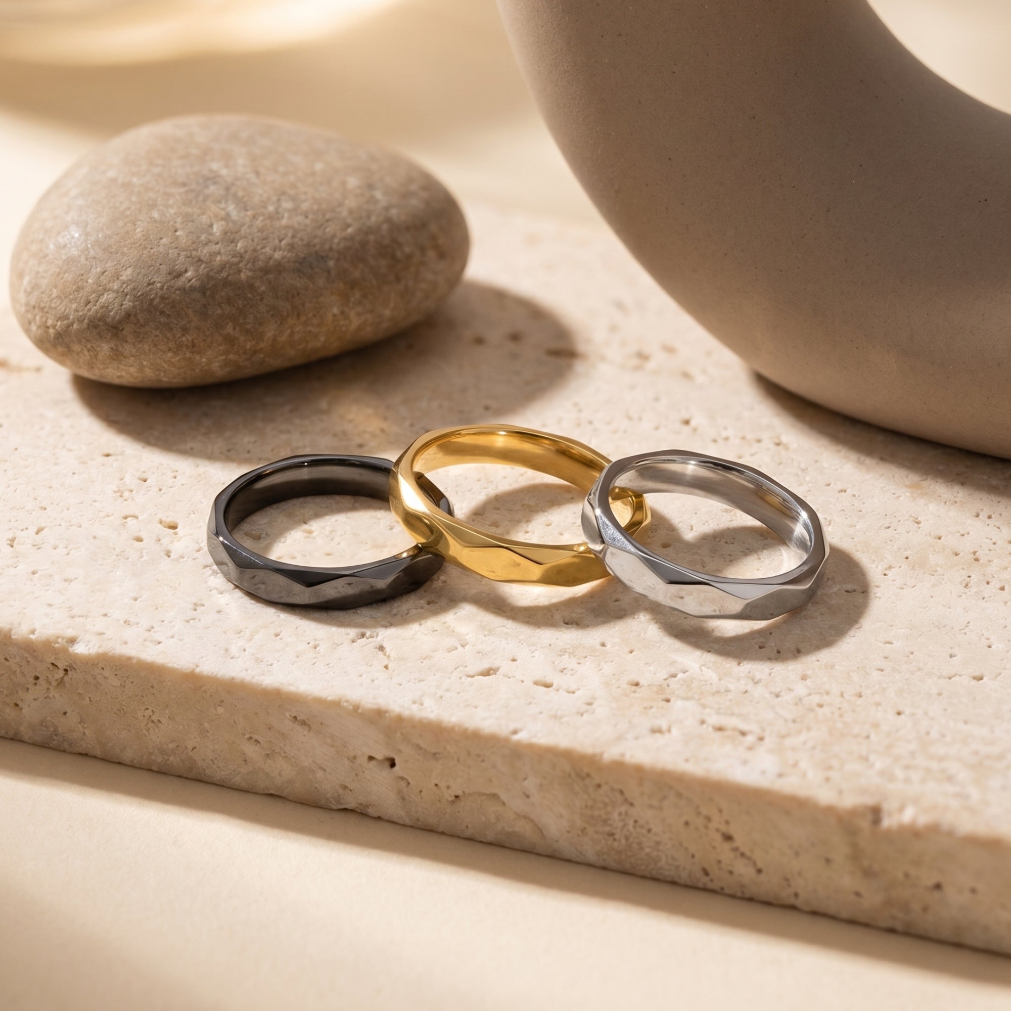 Trio of faceted stainless steel stackable rings in black, gold, and silver on a stone lifestyle background.