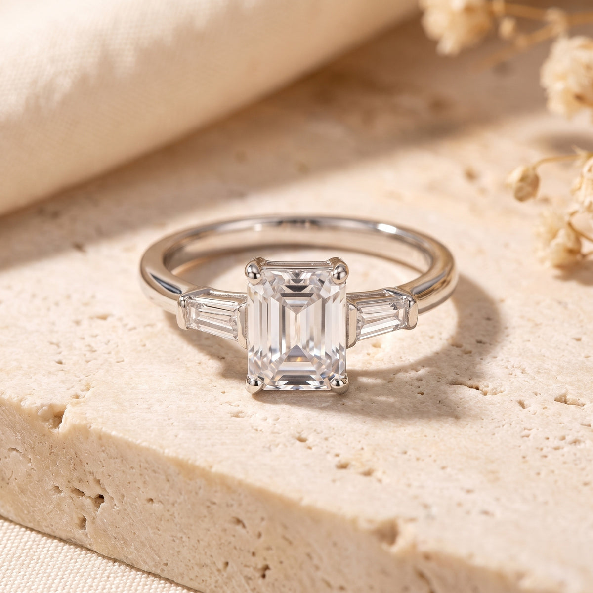 Silver three-stone ring with an emerald-cut center stone and tapered baguette side stones on a beige stone surface.