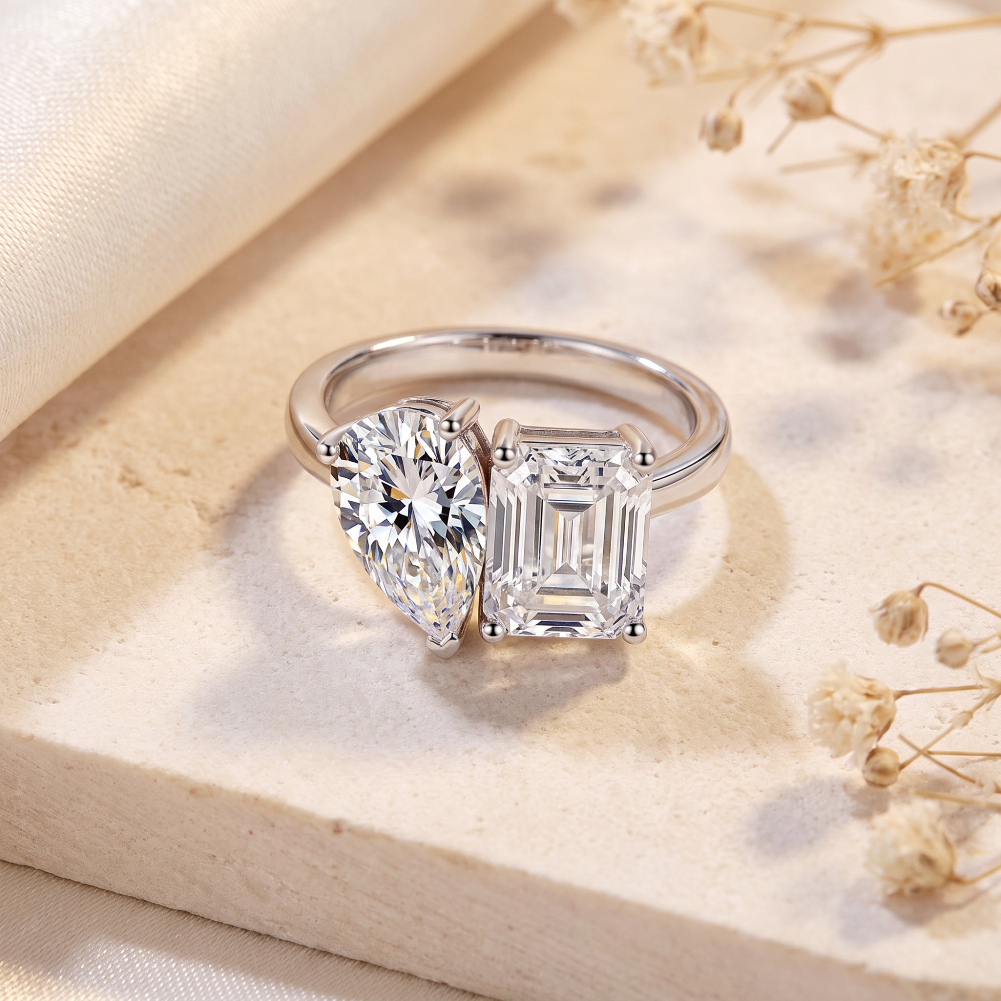 Silver bypass ring with pear and emerald cut simulated diamonds on a stone surface with dried flowers.