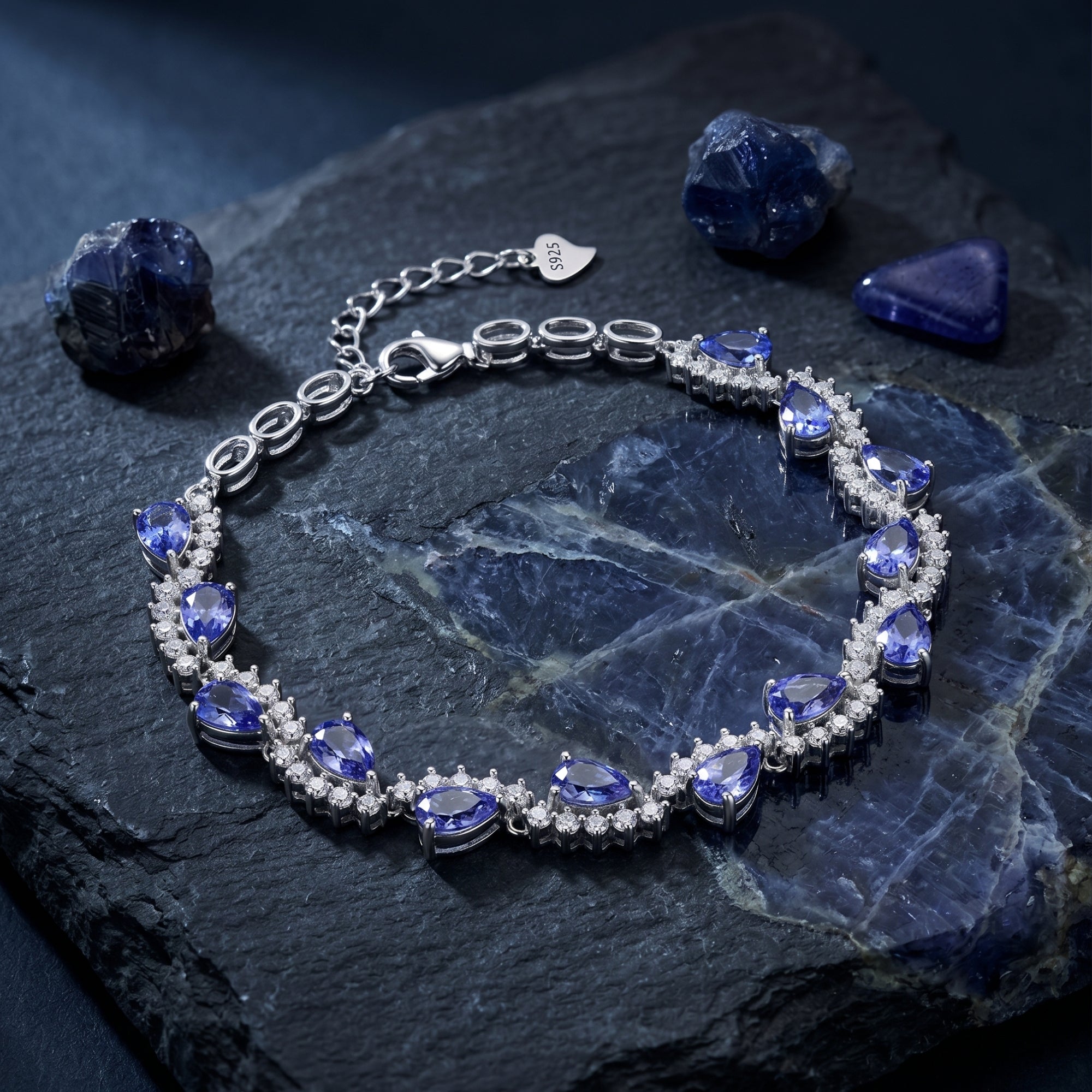 Pear cut tanzanite tennis bracelet in silver displayed on a dark textured stone with raw gemstones.