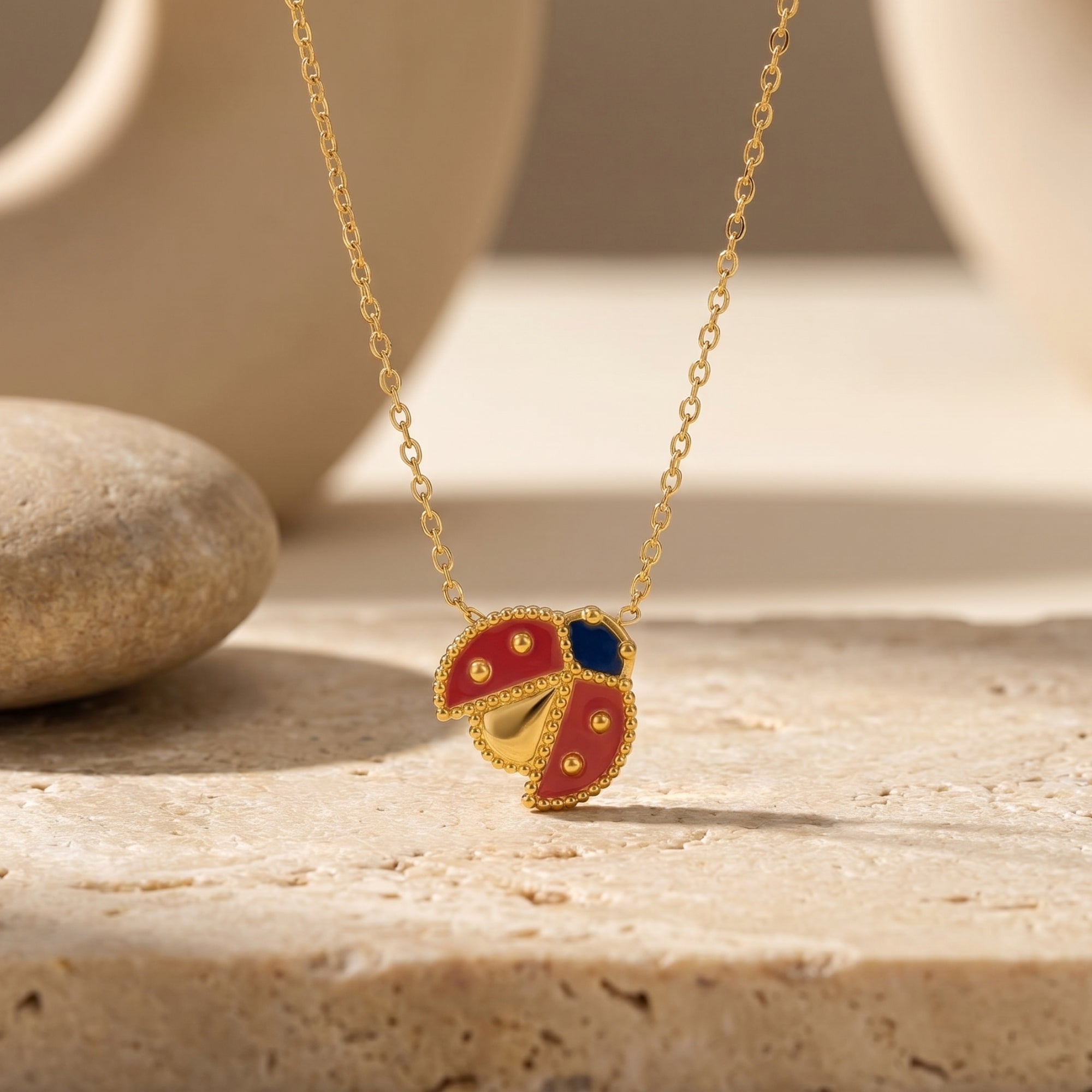 Gold ladybug pendant necklace with red and blue enamel on a stone surface.