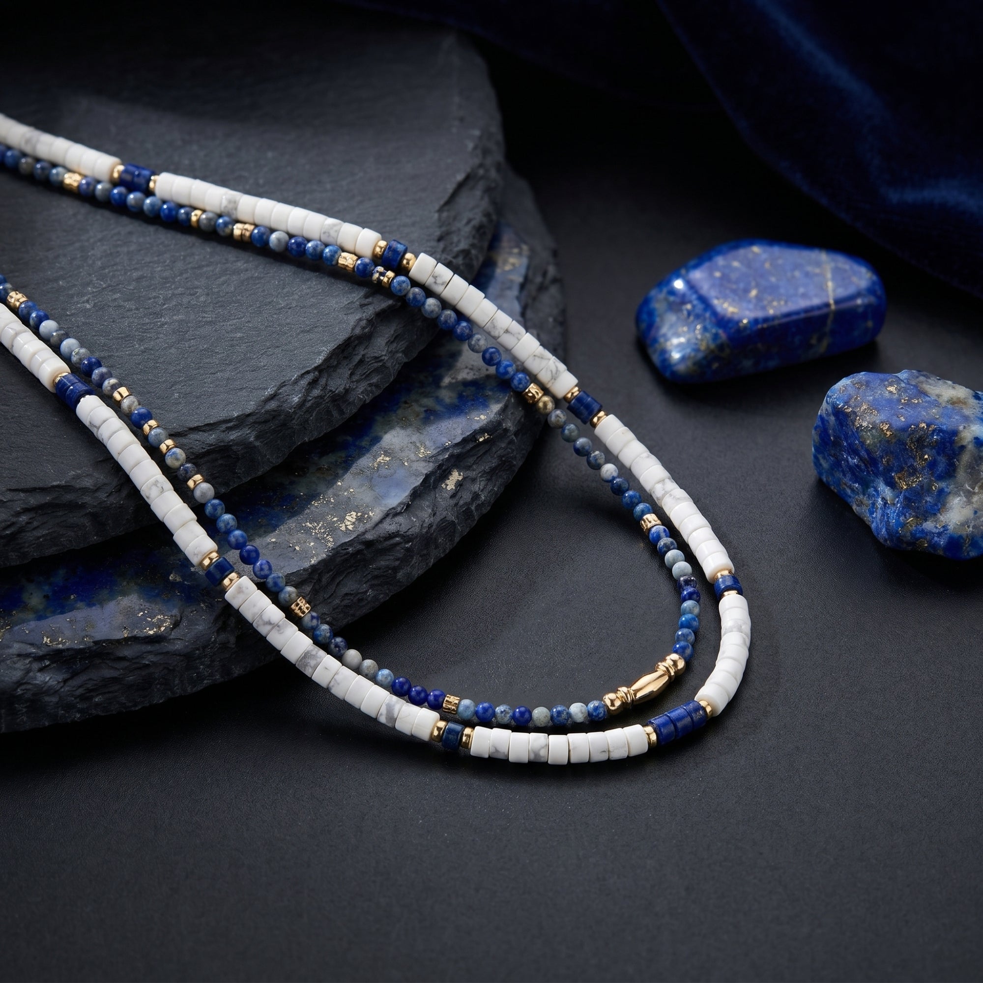 Close-up of layered gold-plated necklaces with round blue lapis and white stone beads on dark slate.