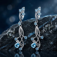 Oceanis Cascade drop earrings with pear and round cut natural blue topaz stones displayed on a dark oceanic rock.