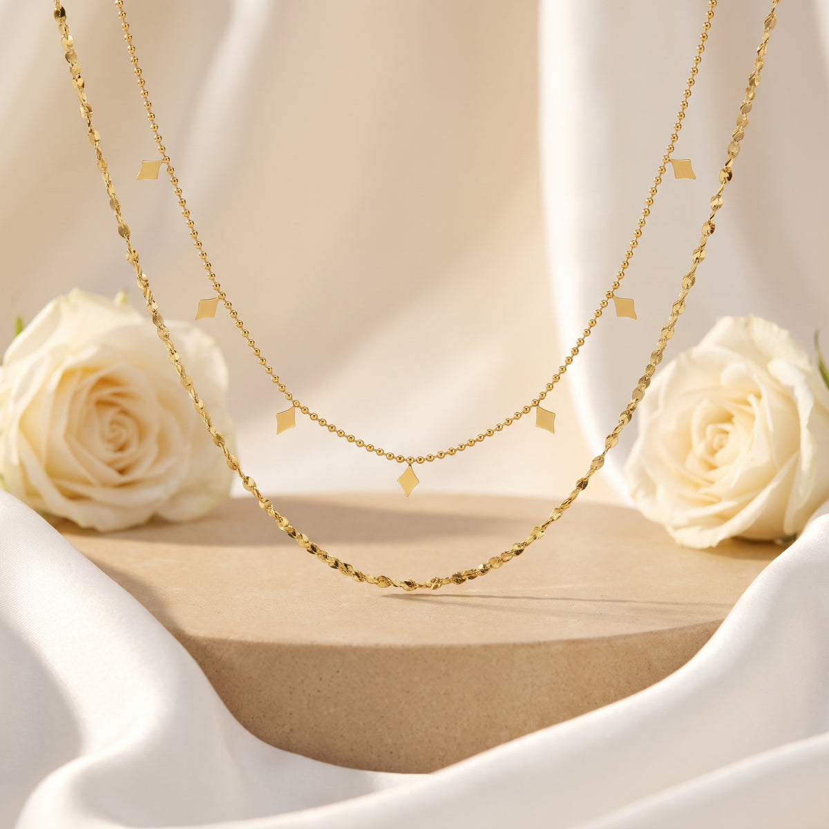 Gold-tone double-strand necklace hanging over a wooden pedestal between two white roses and draped fabric.
