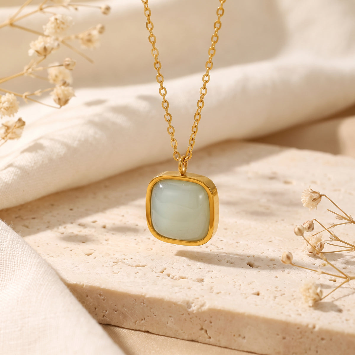 Gold-tone cushion bezel pendant necklace with a pale green smooth center, displayed on a stone slab beside linen.