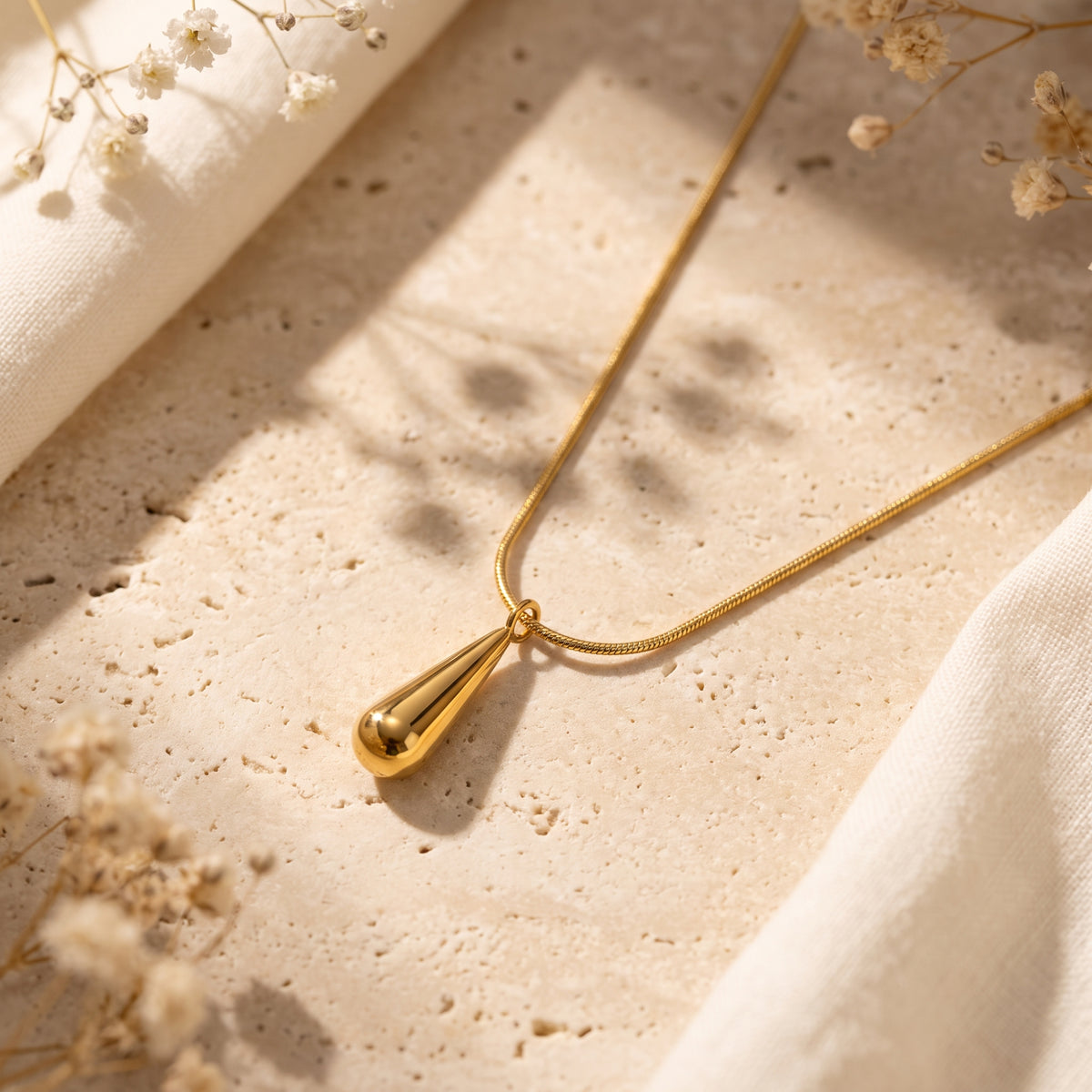 Gold-tone teardrop pendant necklace on a snake chain resting on a stone surface with dried florals.