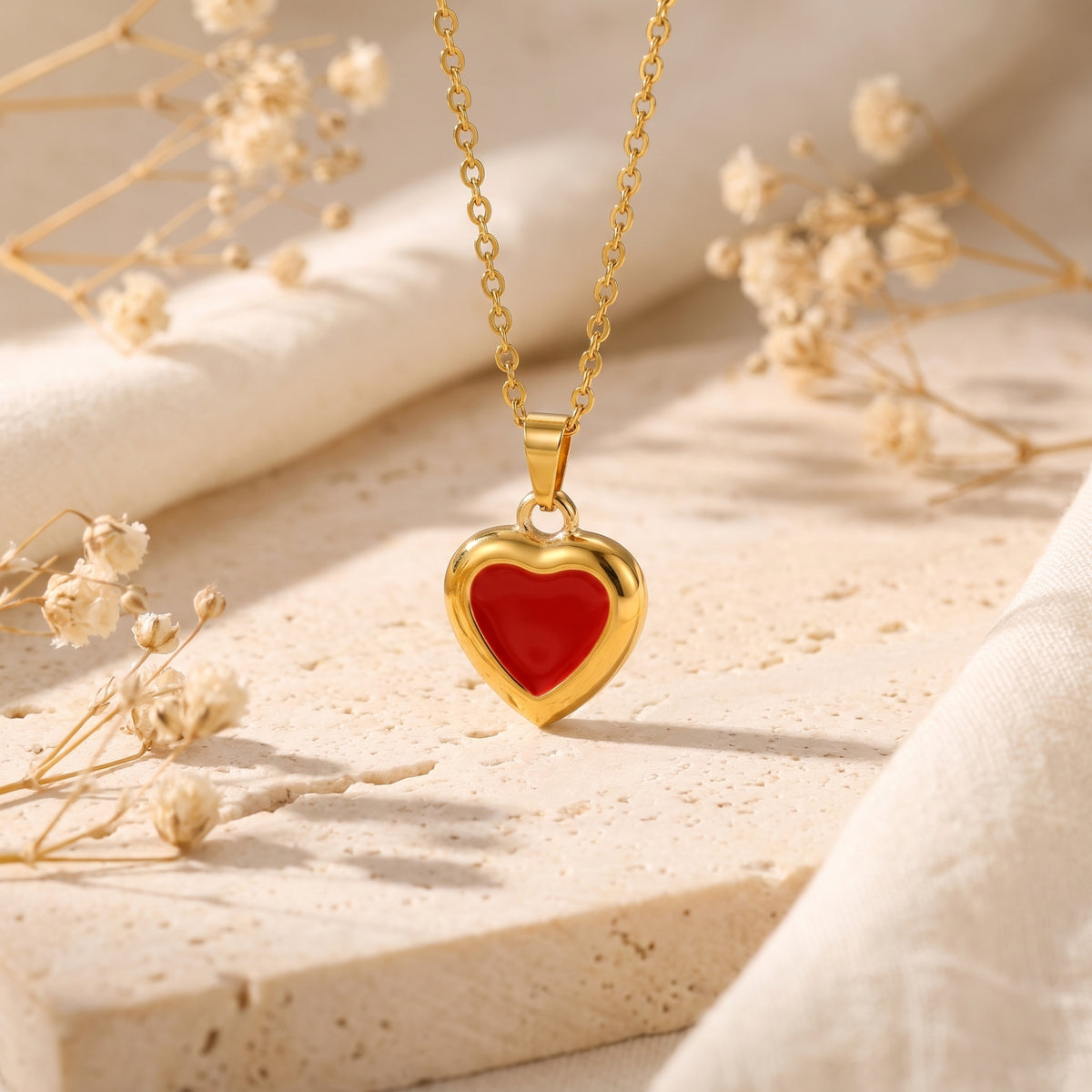 Red enamel heart pendant with a gold-tone frame hanging above a pale stone slab and dried flowers.
