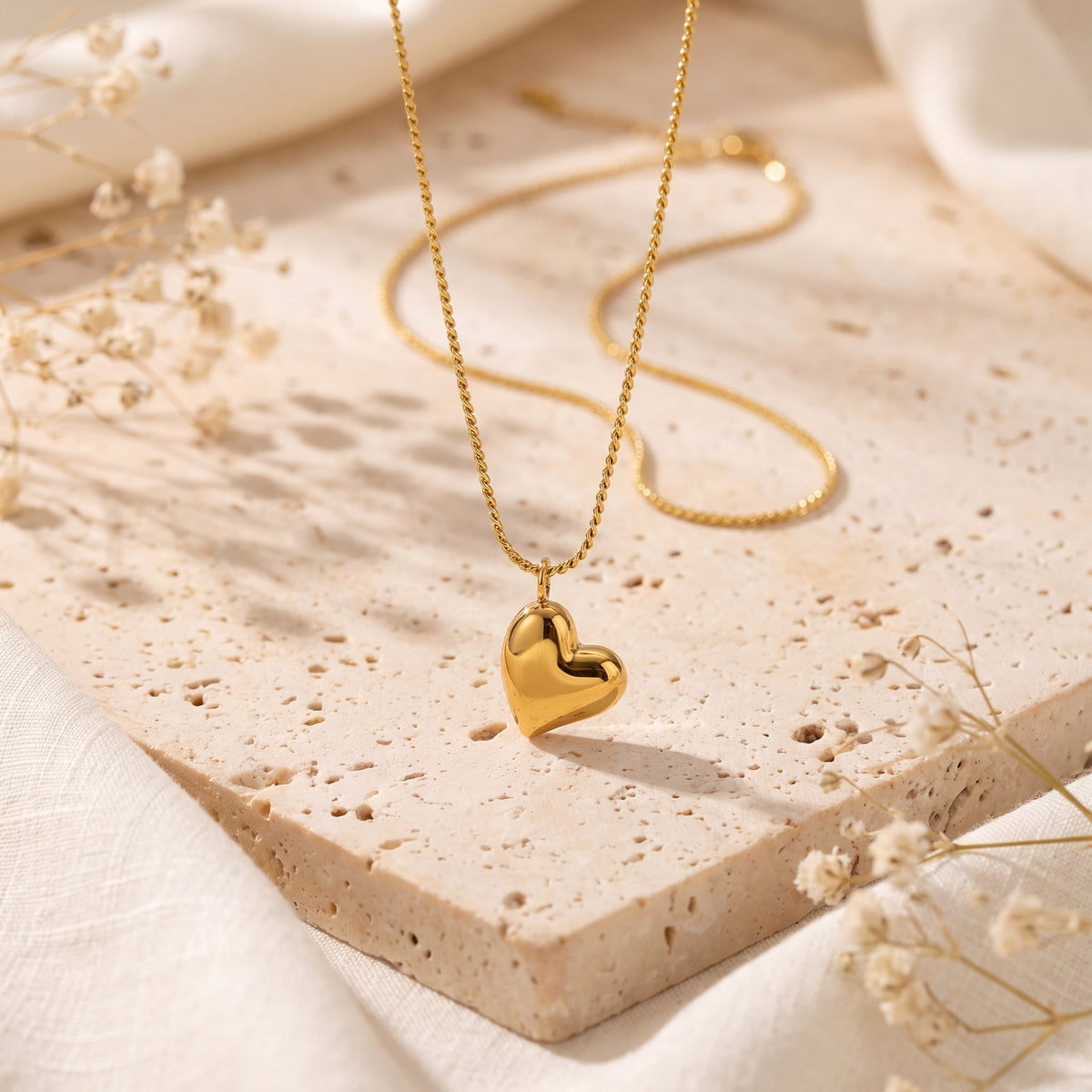 Gold-tone puffed heart pendant necklace displayed over a porous beige stone slab with dried flowers.