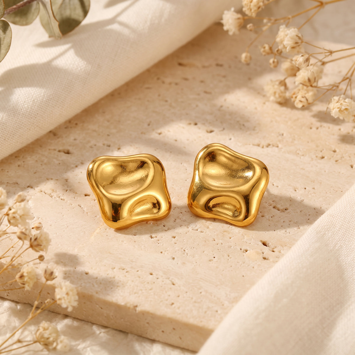 Pair of polished gold-tone sculptural square stud earrings resting side by side on a textured beige stone slab.