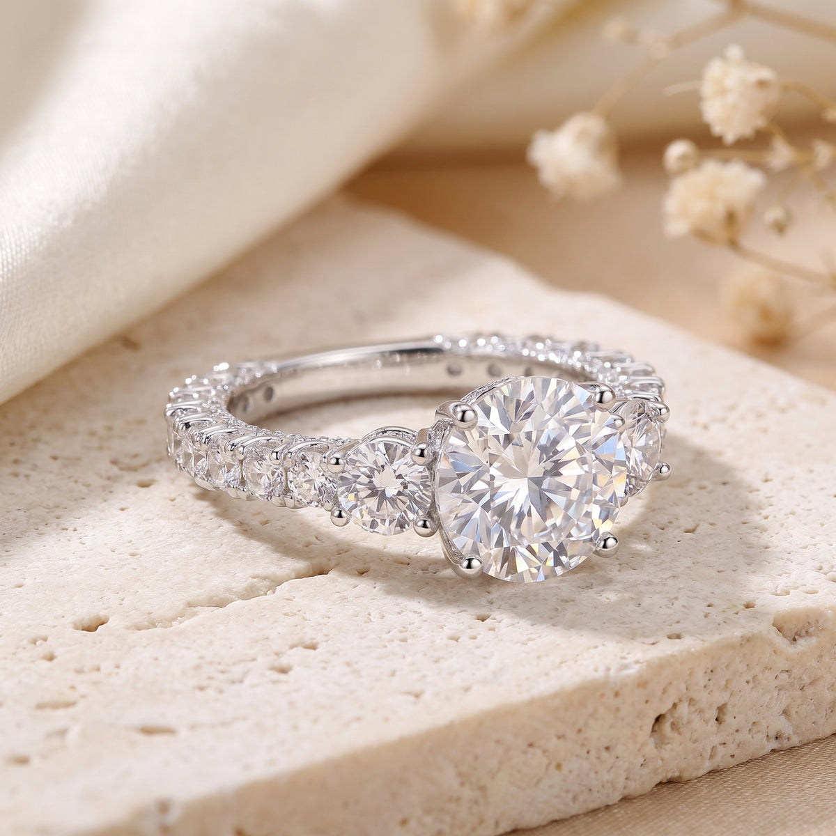 Three-stone silver ring with a large round center gem, side accents, and pave band resting on a beige surface.