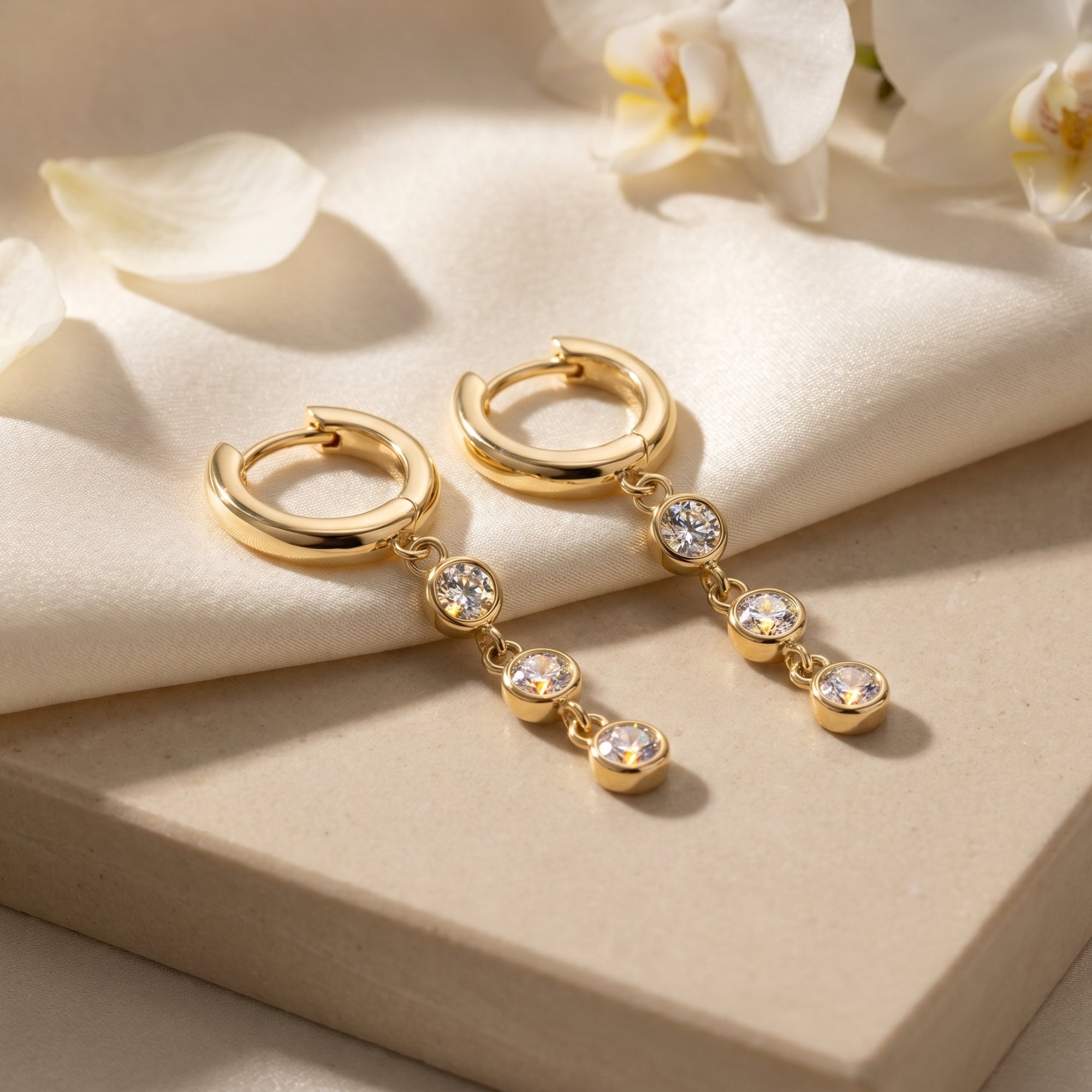 Gold moissanite drop earrings with three round stones on cream fabric and stone with white flowers.