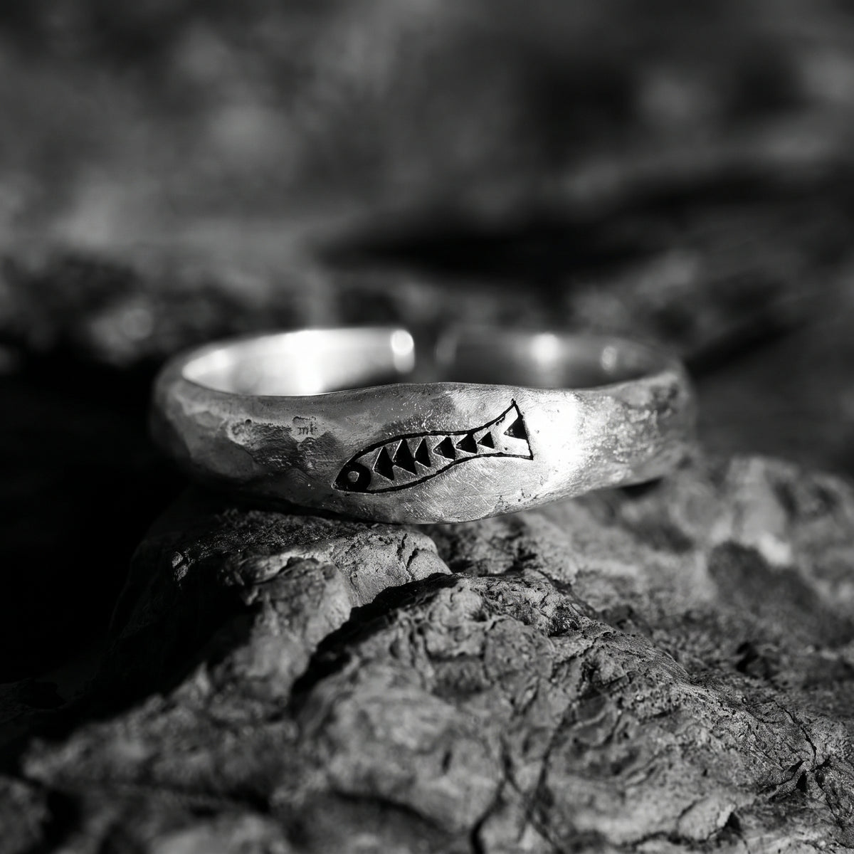 Black-and-white close-up of a silver open ring with a carved fish motif resting on rough stone.