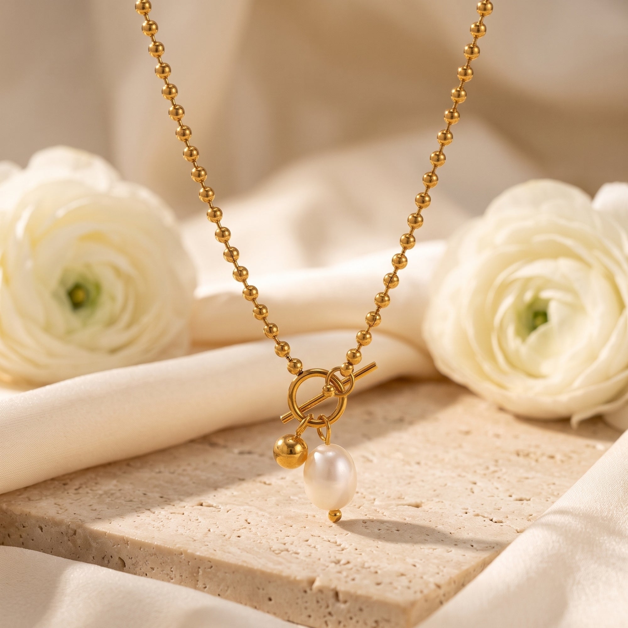 Gold ball chain necklace with baroque pearl and toggle clasp on a styled stone background.