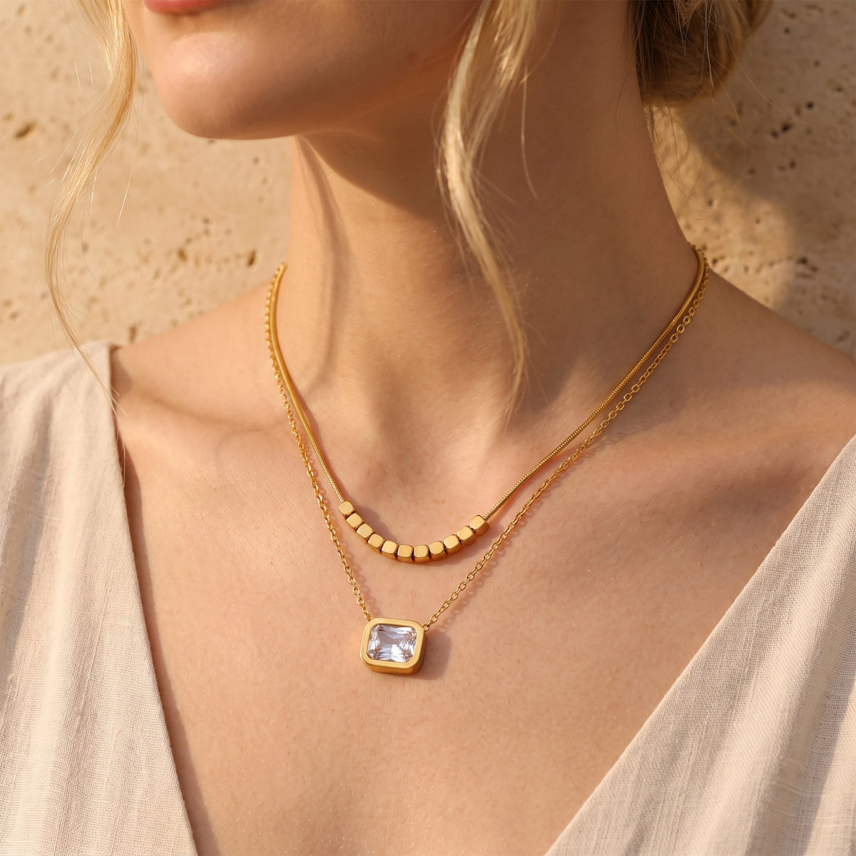 Model wearing a gold-tone double-layered necklace with a cube-beaded chain and square clear pendant.