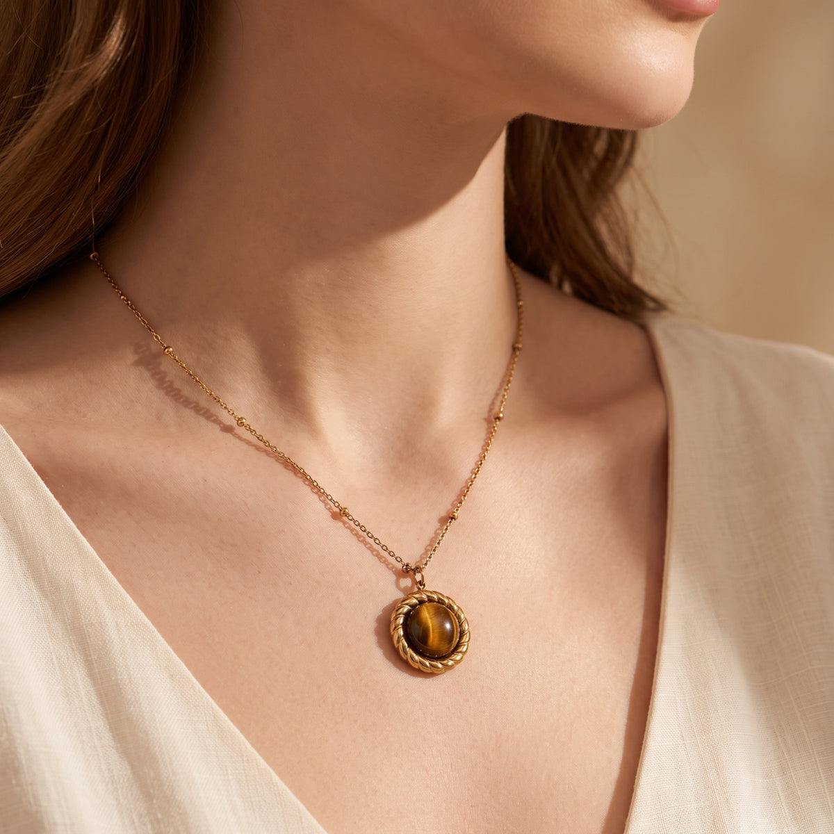 Side view of a model wearing a gold-tone station chain with a round brown cabochon pendant over a white top