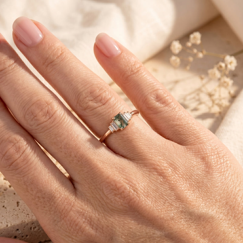 Rose gold emerald-cut moss agate ring with baguette side stones worn on a woman's finger.