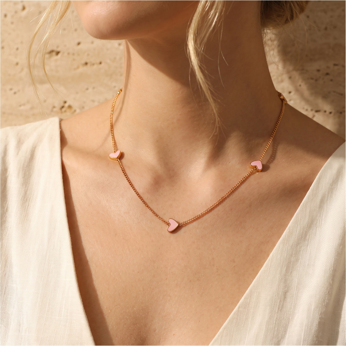 Gold-tone station necklace with small pink enamel hearts worn along the collarbone with a white V-neck top.