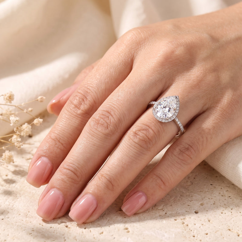 Hand wearing a silver pear-cut halo ring against a warm, softly textured fabric background.