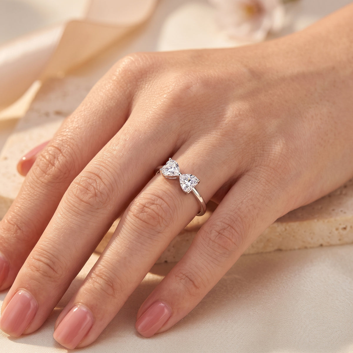 Close-up of the silver bow ring with twin heart-shaped clear stones worn on a hand with pink nails.