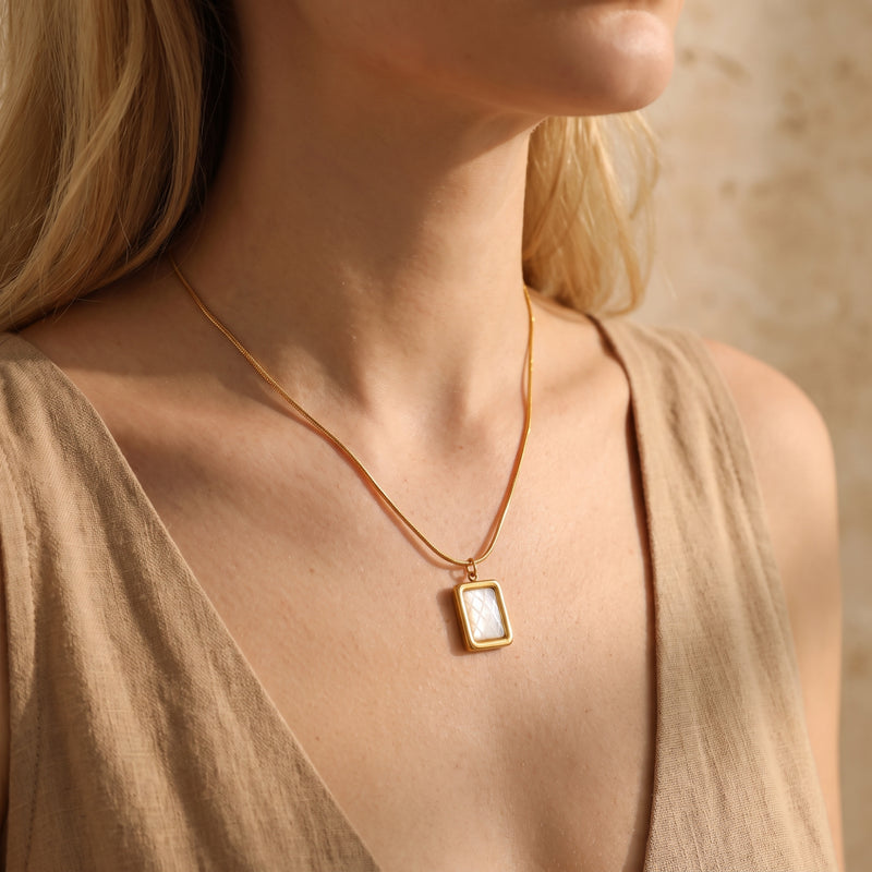 Gold-tone necklace with a rectangular quilted pendant worn around a woman's neck over a beige top.