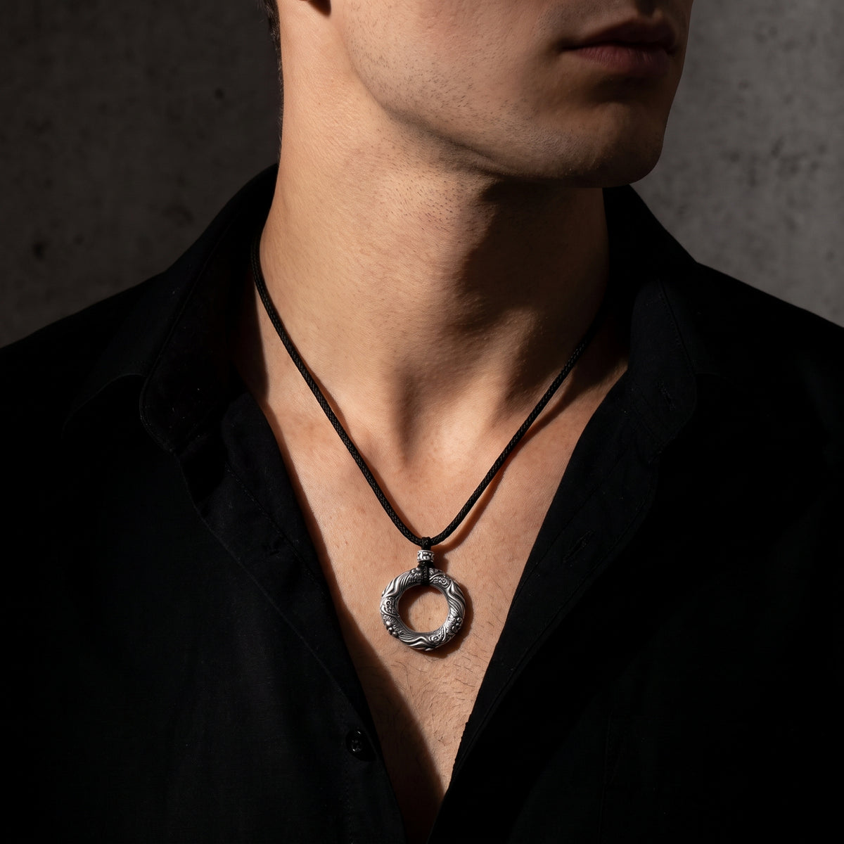 Man wearing the carved circular silver pendant necklace with an unbuttoned black collared shirt.