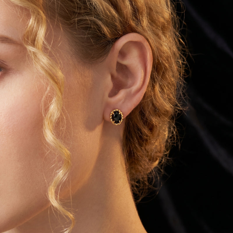 Model wearing a natural black onyx oval stud earring with an 18K gold plated twisted border.