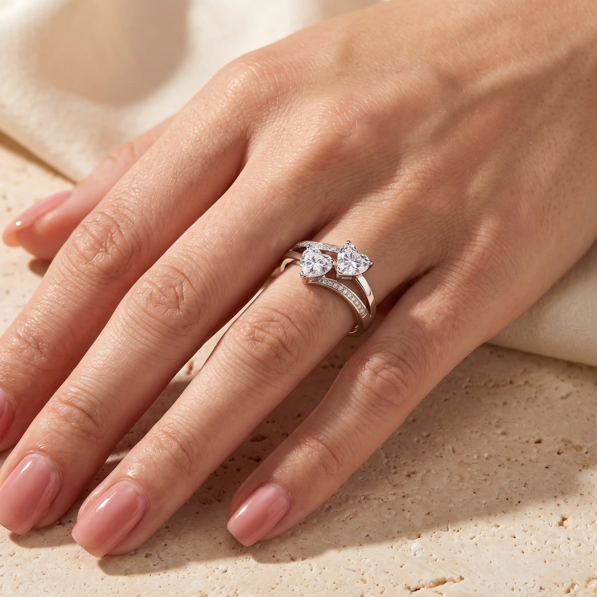 Silver twin heart bypass ring with clear heart-shaped stones and pavé accents worn on a finger.