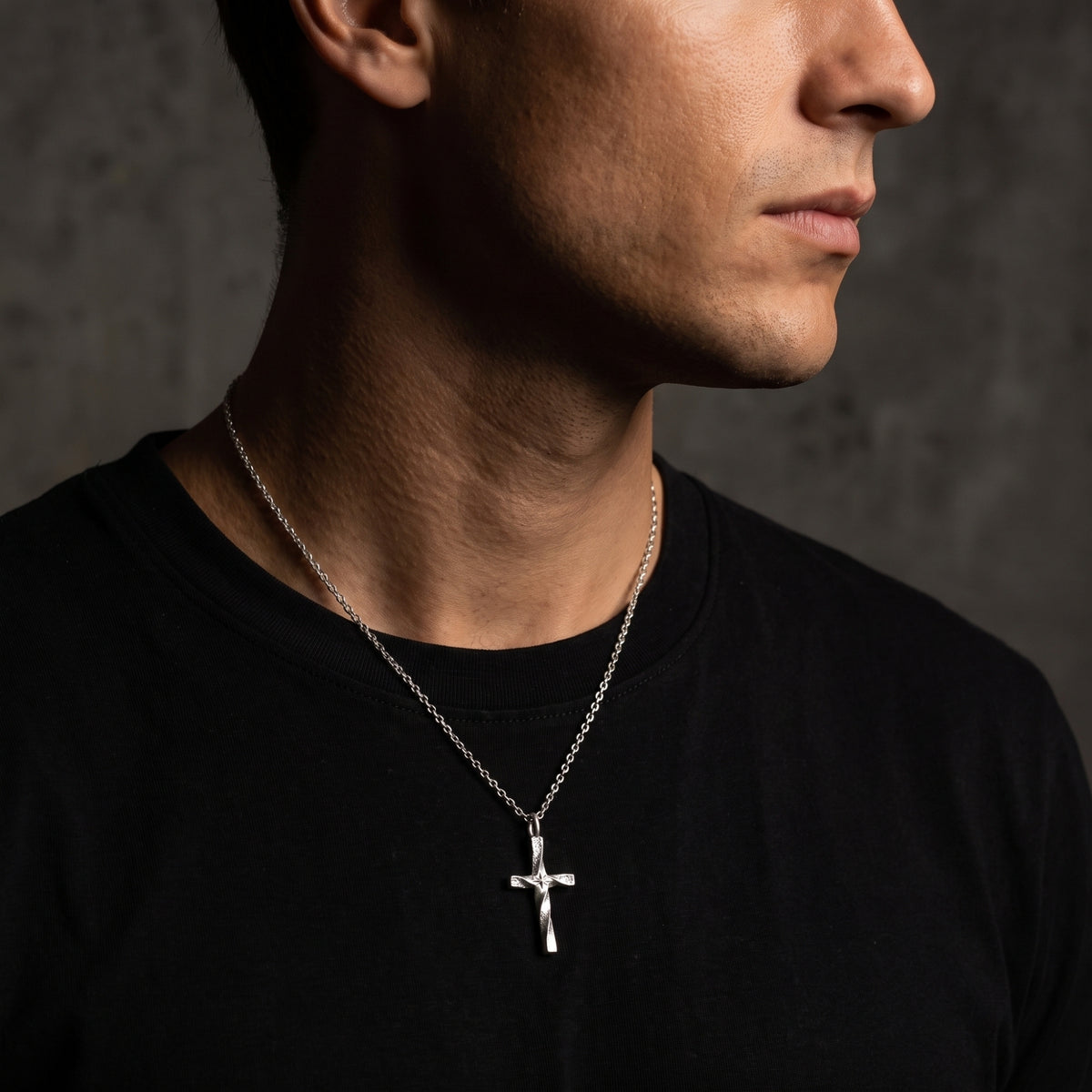Silver twisted cross necklace worn by a man in profile over a black T-shirt.