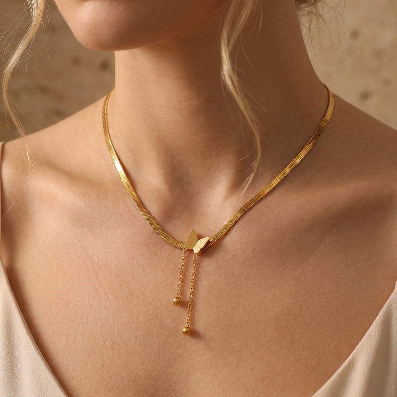 Model wearing a gold-tone flat chain necklace with a butterfly pendant and two dangling ball-ended chains.