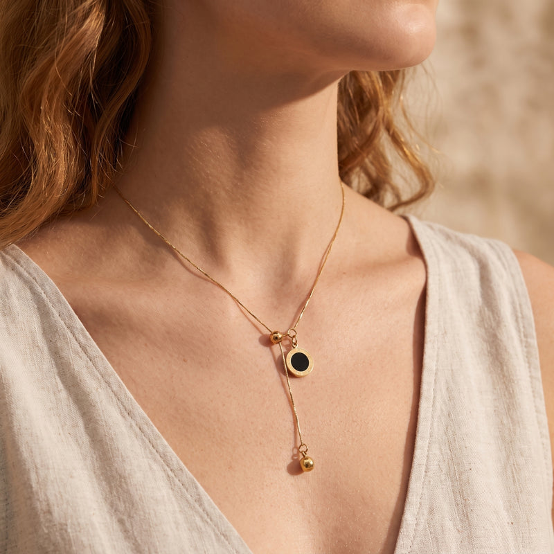 Gold-tone lariat necklace with black Roman numeral disc pendant worn on a model in a light beige sleeveless top.