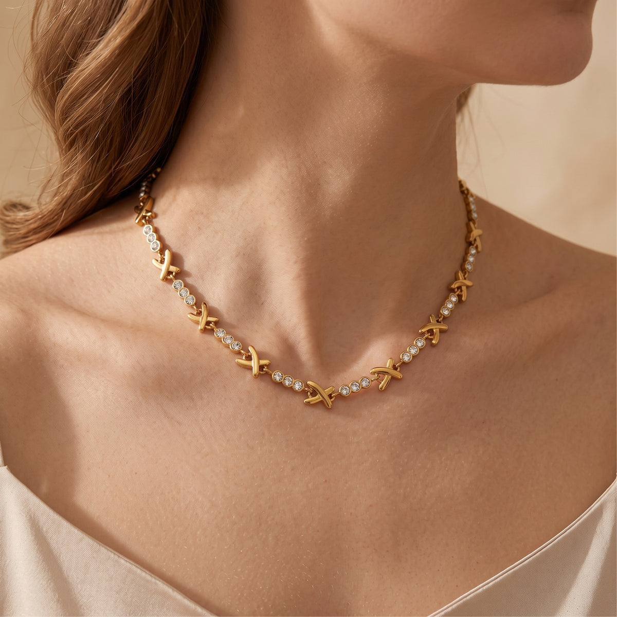 Gold-tone X station necklace worn around the neck of a model wearing a neutral silk top.