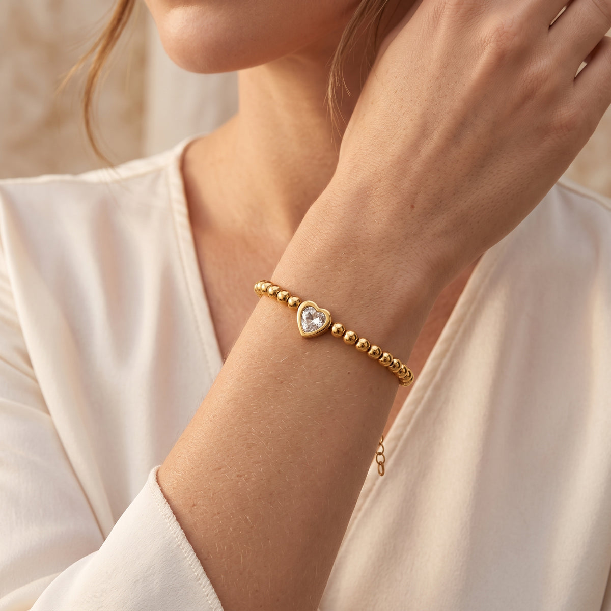 Gold-tone beaded bracelet worn on a wrist, with a heart-shaped clear stone centered over a white sleeve.