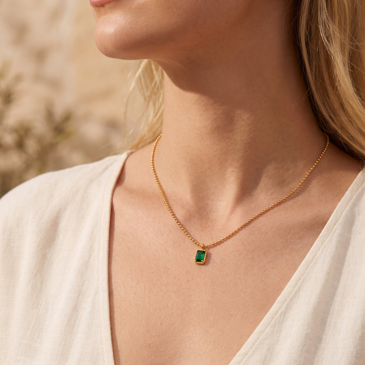 Side profile of a model wearing the gold-tone ball chain necklace with a green emerald-cut pendant.