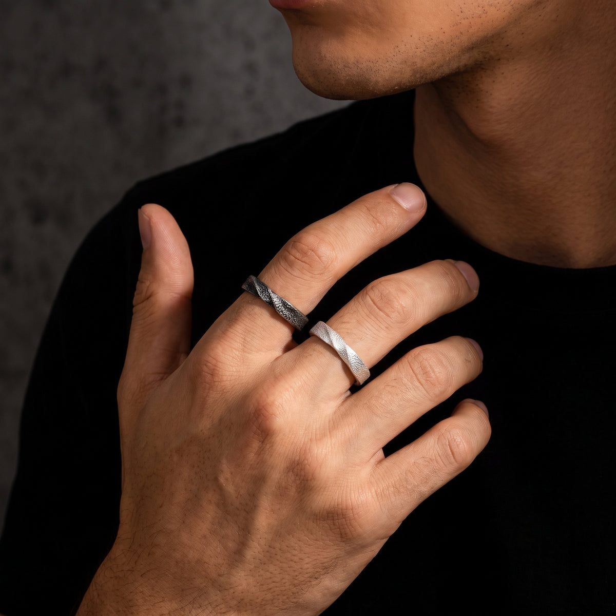 Man wearing dark and light textured open-band rings on his index and middle fingers.