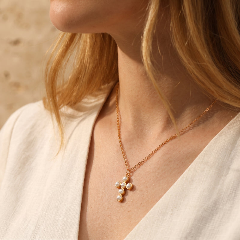 Angled lifestyle view of a woman wearing the gold chain necklace with a pearl cross pendant over a cream top.