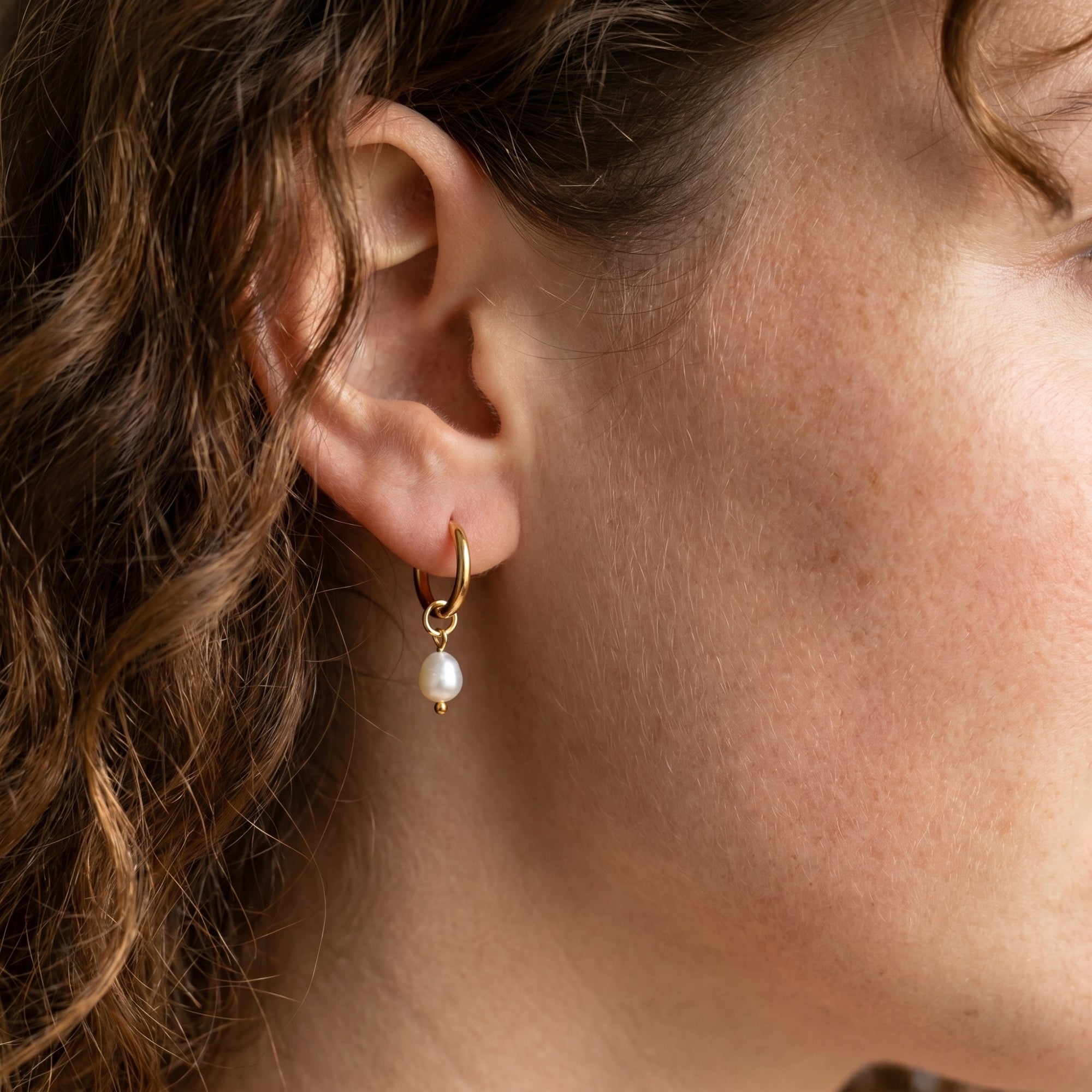 Gold hoop earring with oval pearl worn on ear.
