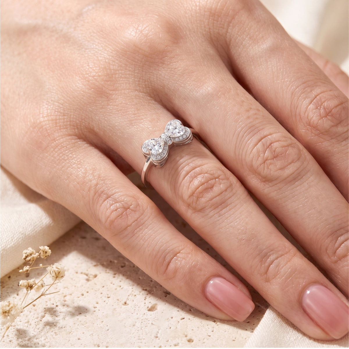 Silver bow ring with twin heart-cut halo stones worn on a hand’s middle finger.