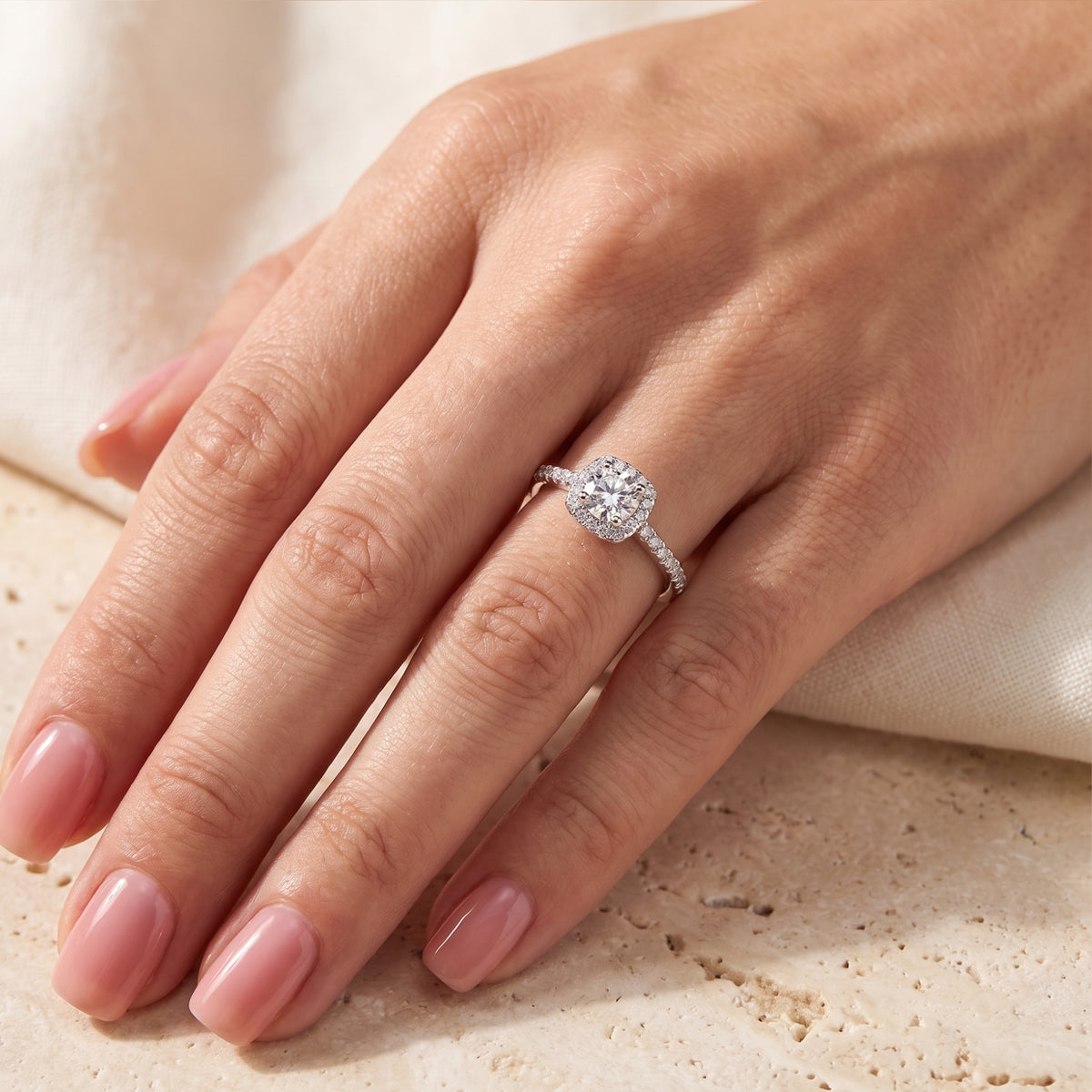The silver cushion halo ring worn on a hand, viewed from above against cream fabric.
