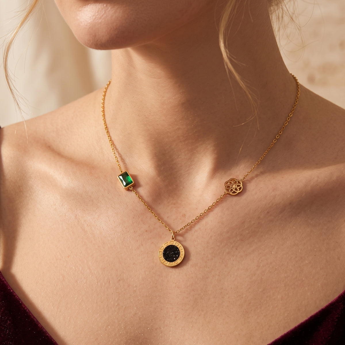 Model wearing a gold-tone station necklace with a green crystal, floral wheel, and black medallion over a burgundy top.