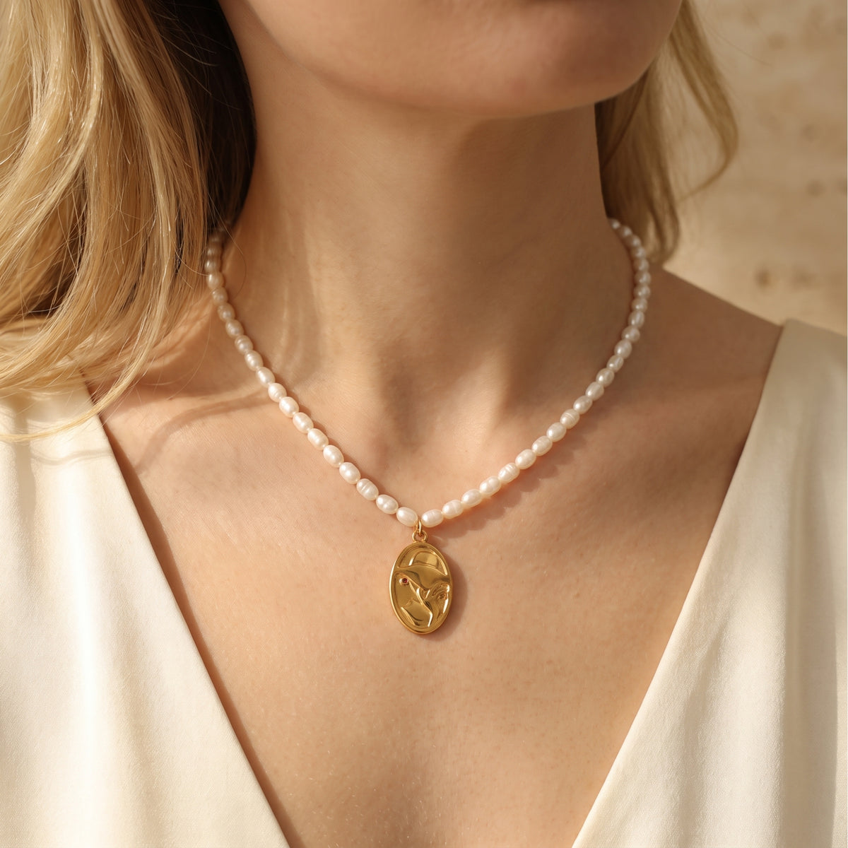 Close-up of a gold-tone oval face-profile pendant with a small red accent, worn on a white pearl necklace.