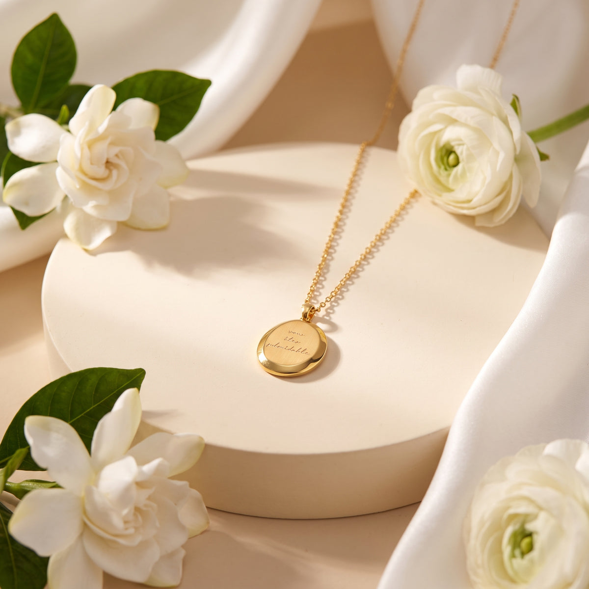 Gold-tone round medallion necklace with cursive engraving on a cream pedestal surrounded by white flowers and green leaves.