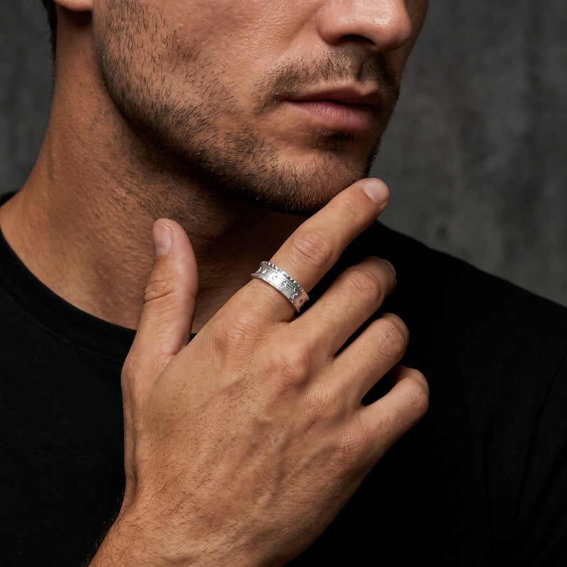 Man wearing a wide silver ring with raised motifs on his index finger while touching his chin.