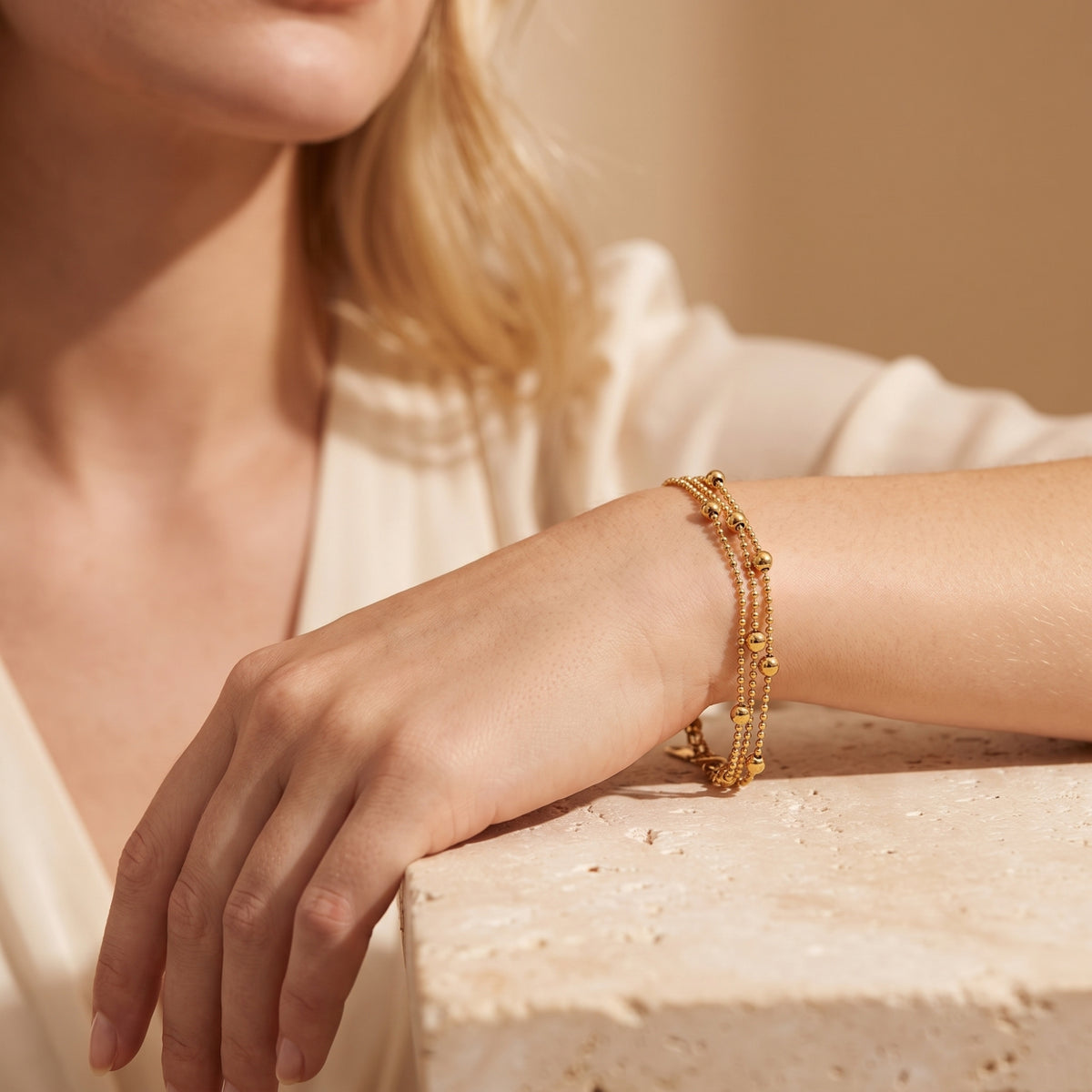 Gold-tone three-strand bead bracelet worn on a wrist over a textured light-colored block.